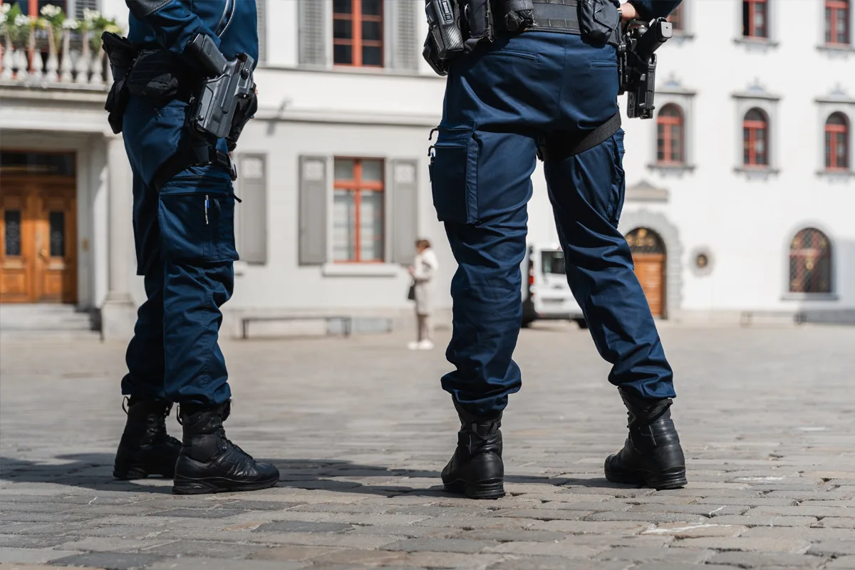 Zwei Polizisten in blauer Uniform sind von der Hüfte abwärts zu sehen, während sie auf einem Kopfsteinpflasterplatz vor einem alten Gebäude stehen.