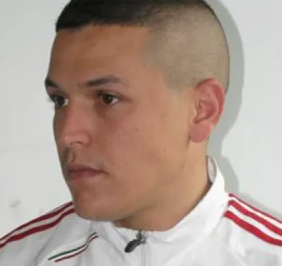 Ein Mann mit kurzem Haar, frontal fotografiert, trägt einen weißen Trainingsanzug mit roten Streifen. Der Hintergrund ist neutral.