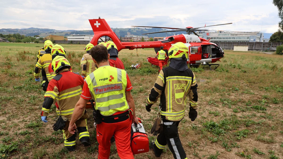 Mehrere Rettungskräfte, teils in Feuerwehruniformen, nähern sich einem Rega-Helikopter auf einer Wiese. Im Hintergrund sind Gebäude zu erkennen.