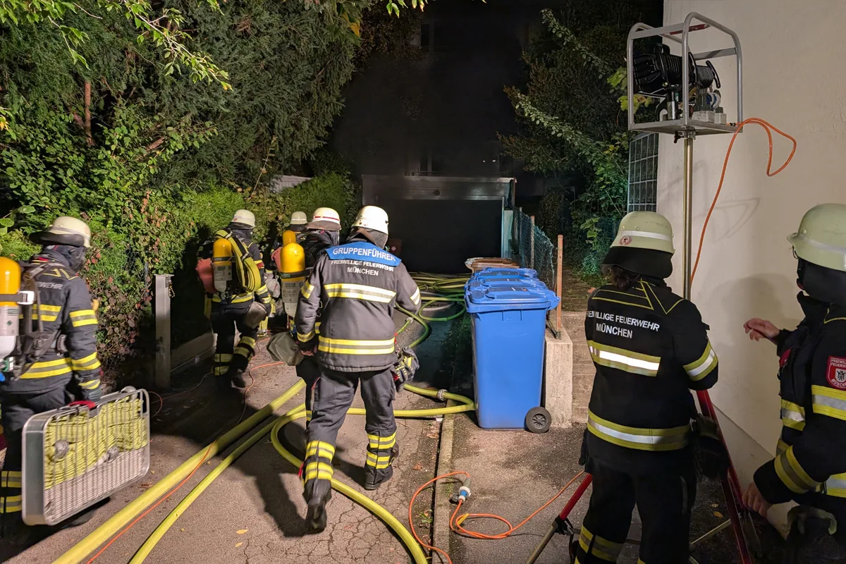 Mehrere Feuerwehrleute in Schutzkleidung und Helmen arbeiten vor einem dunklen Gebäude. Schläuche und Beleuchtungsausrüstung sind sichtbar.