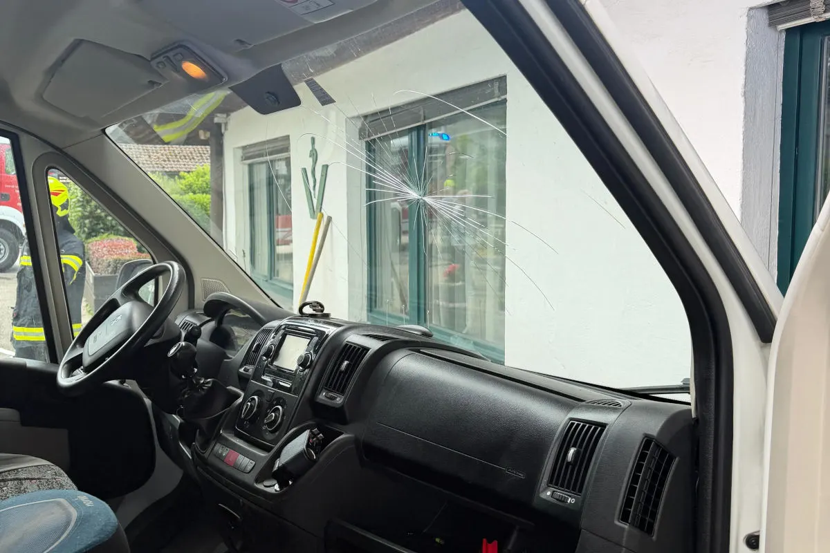 Im Vordergrund sieht man die Innenansicht eines Fahrzeugs mit einem Riss in der Windschutzscheibe. Im Hintergrund stehen ein Feuerwehrmann in Uniform und ein weiteres Fahrzeug.