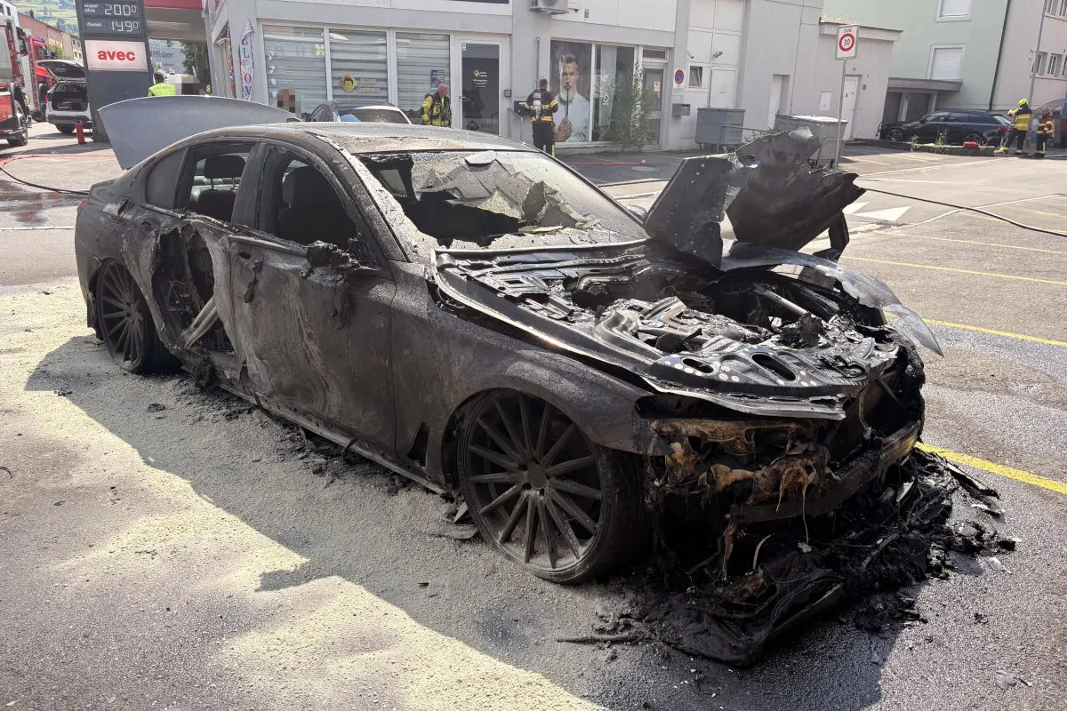 Ein schwarzer Pkw mit schweren Brandschäden steht auf einem Parkplatz, umgeben von Löschmitteln. Feuerwehrleute sind im Hintergrund erkennbar.
