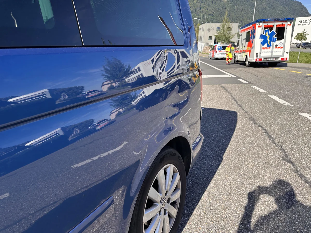 Nahaufnahme eines blauen Autos mit Beule auf einer Straße. Im Hintergrund ein Rettungswagen und eine Person in leuchtender Kleidung.