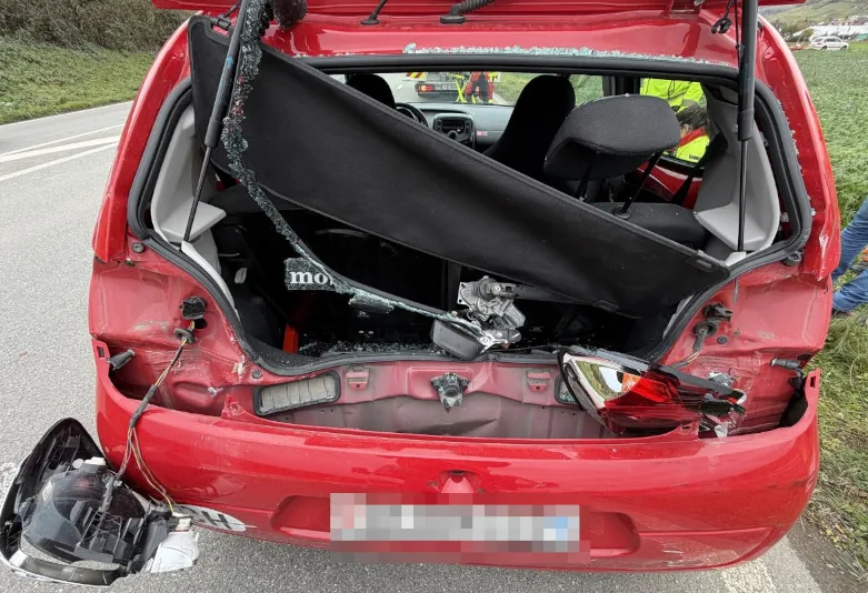 Das Bild zeigt das stark beschädigte Heck eines roten Autos auf einer Landstraße. Hinter- und Rücklichter sind zerstört, und im Innenraum ist die Rücksitzbank sichtbar.