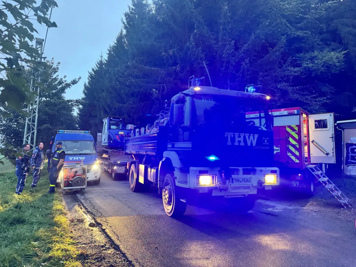 Zwei THW-Fahrzeuge stehen auf einer Straße am Wald, während Einsatzkräfte neben ihnen stehen. Es herrscht Dämmerung oder frühe Morgenstunden mit blauem Lichtschein.