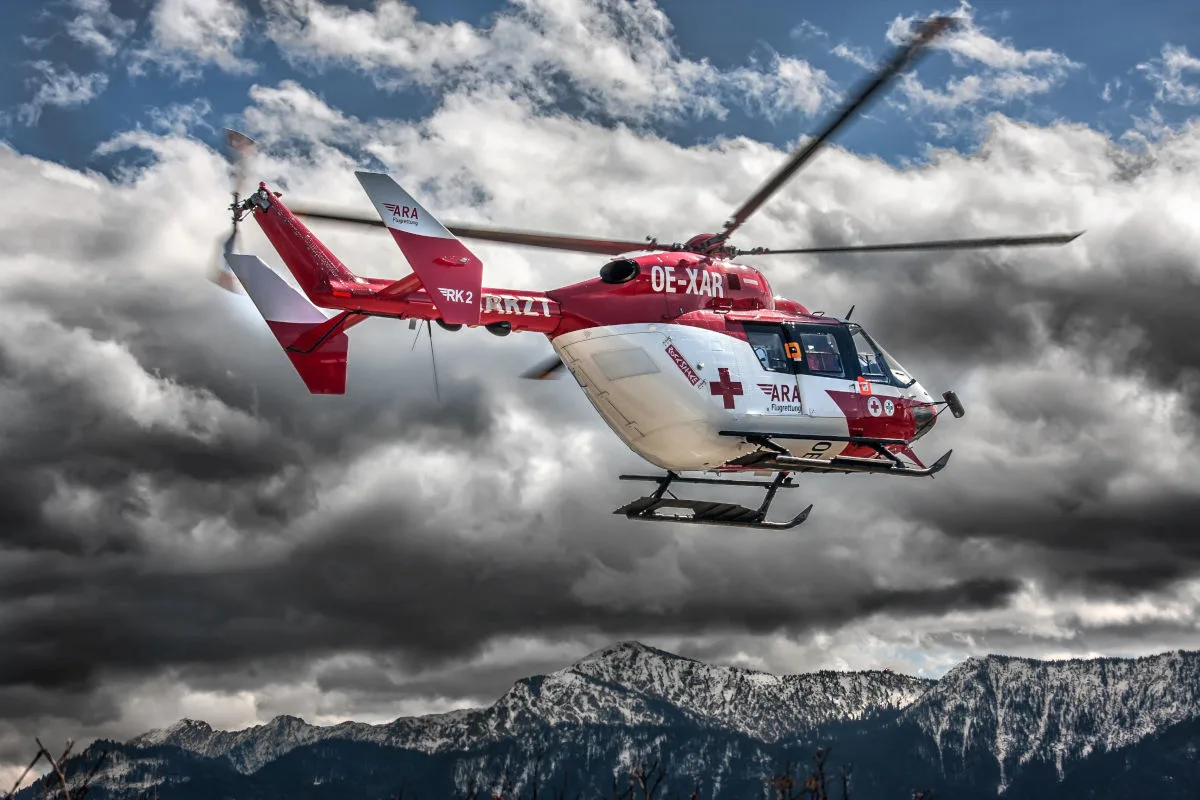 Ein roter Rettungshubschrauber der ARA Flugrettung fliegt über eine verschneite Berglandschaft im Hintergrund. Der Himmel ist bewölkt.