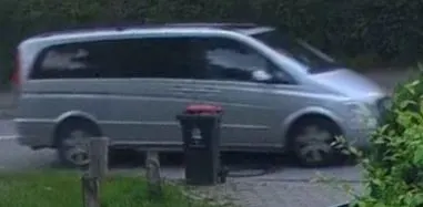 Ein silberner Van fährt schnell auf einer Straße vorbei, neben einer Mülltonne und Gebüsch, die den Vordergrund bilden.