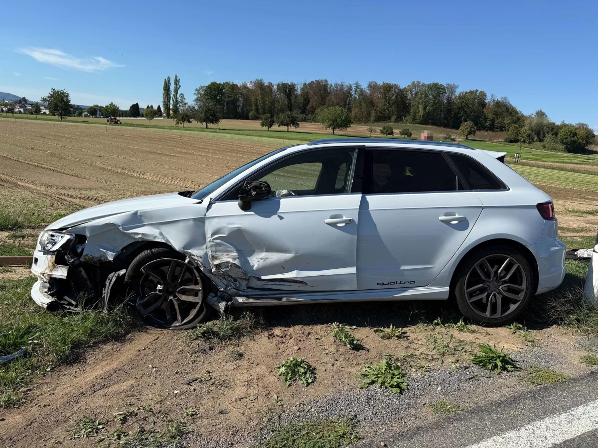Ein weißer Audi mit erheblichen Beschädigungen an der linken Seite steht auf einem Grünstreifen neben einer Landstraße bei klarem Himmel.