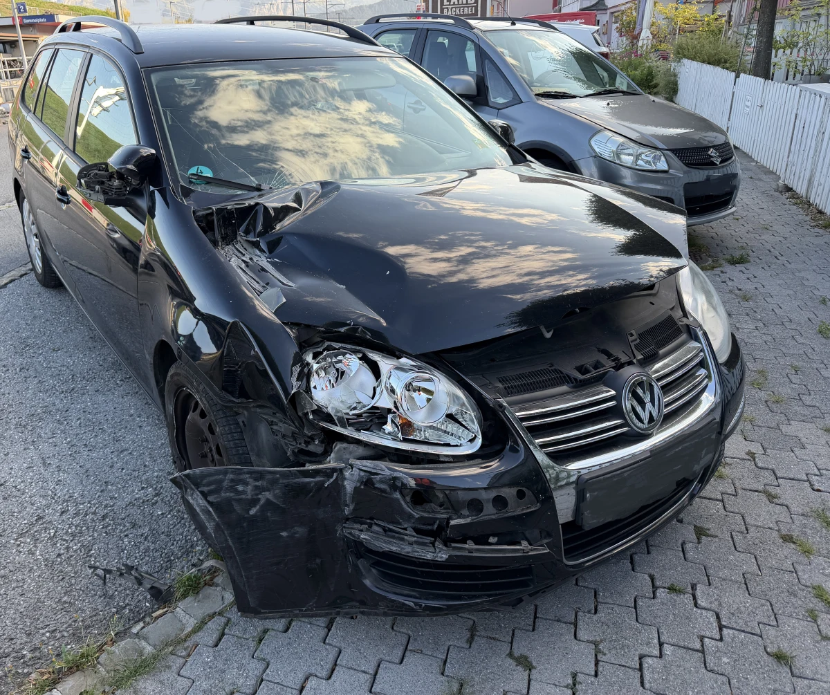 Ein schwarzer VW mit deutlichen Schadenspuren an der Front steht auf einem gepflasterten Parkplatz. Der linke Frontbereich und Scheinwerfer sind stark deformiert.