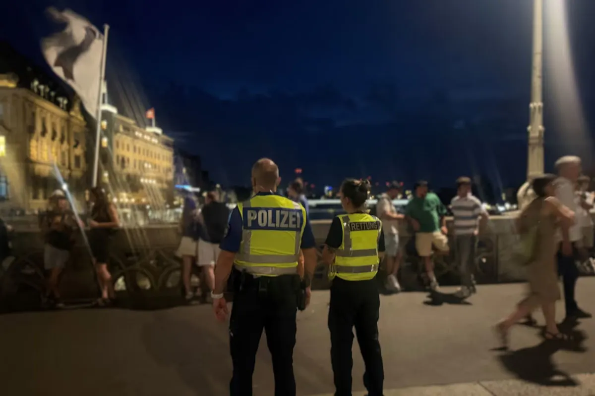 Ein Polizist und eine Rettungskraft in Warnwesten stehen abends auf einer Brücke, umgeben von einer Menschenmenge. Im Hintergrund sind beleuchtete Gebäude und ein dunkler Himmel sichtbar.