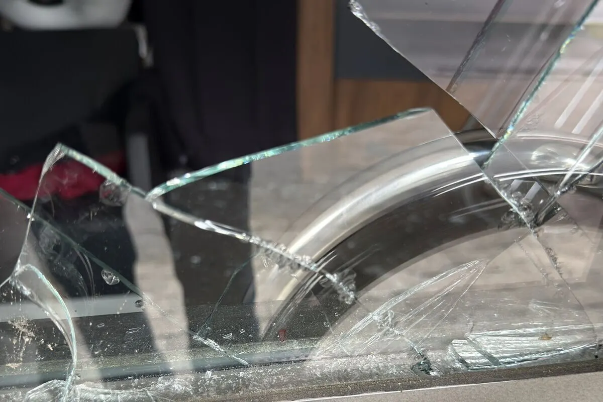 Das Bild zeigt eine zerbrochene Glasscheibe, deren Scherben in verschiedene Richtungen auf einer Oberfläche verteilt sind. Im Hintergrund sind Teile eines weiteren Objekts, möglicherweise eines Fahrzeugs, sichtbar.