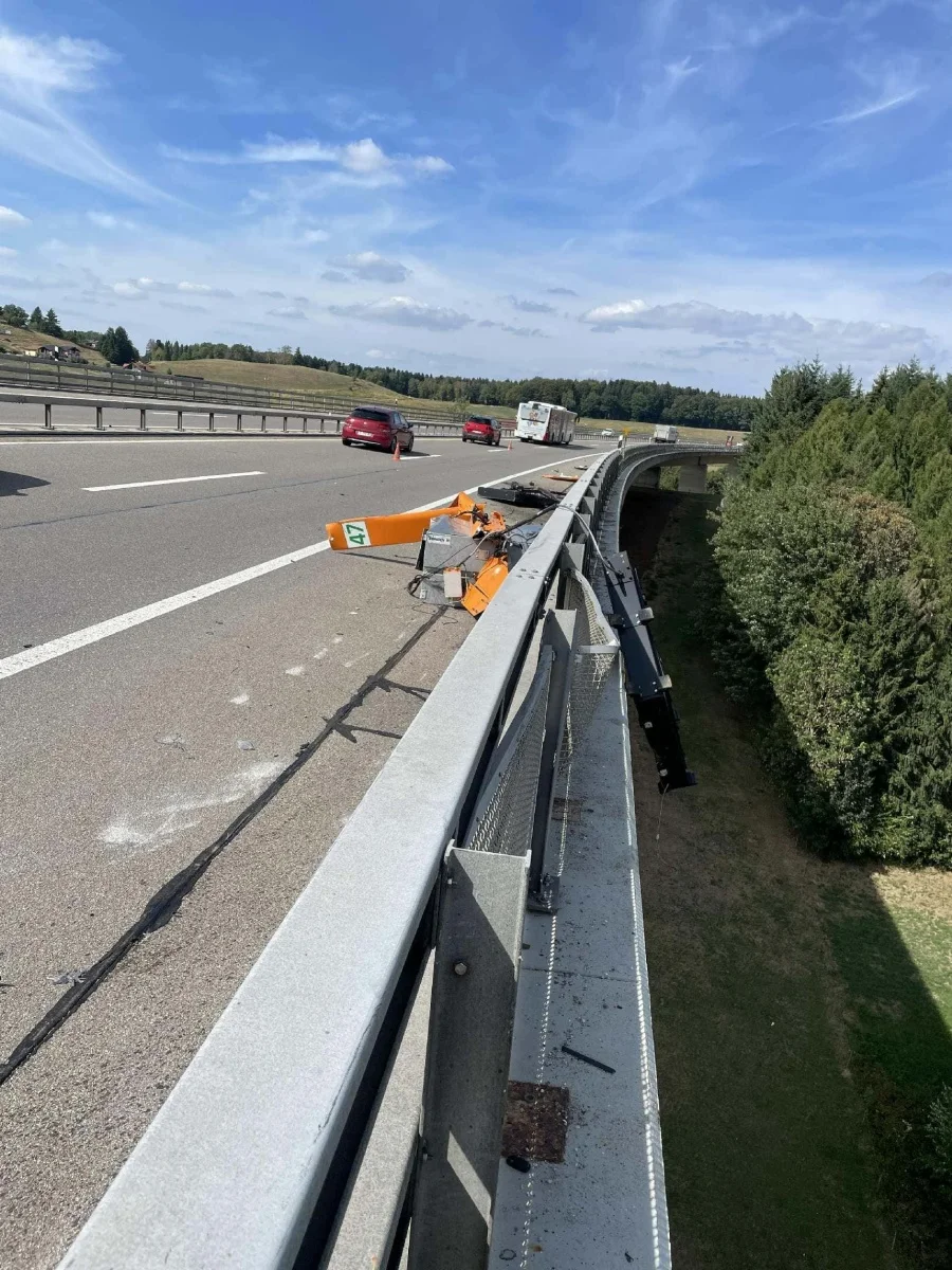 Auf einer Autobahn ist eine Leitplanke stark beschädigt, während Autos die Unfallstelle passieren. Der Himmel ist klar und es ist tagsüber.