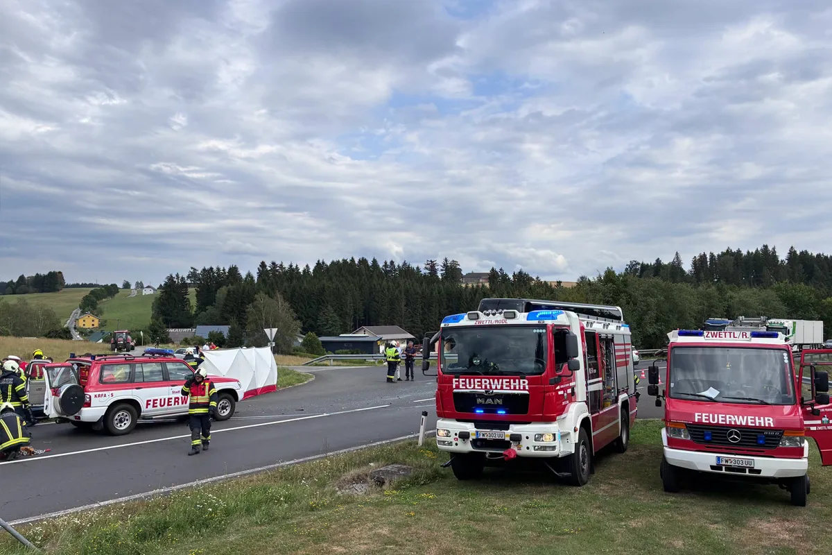 Mehrere Feuerwehrfahrzeuge und Einsatzkräfte befinden sich an einem Straßenrand neben einer Unfallstelle. Ein weißes Zelt verdeckt teilweise die Unfallstelle, während Einsatzkräfte in Schutzkleidung sich in der Nähe aufhalten.