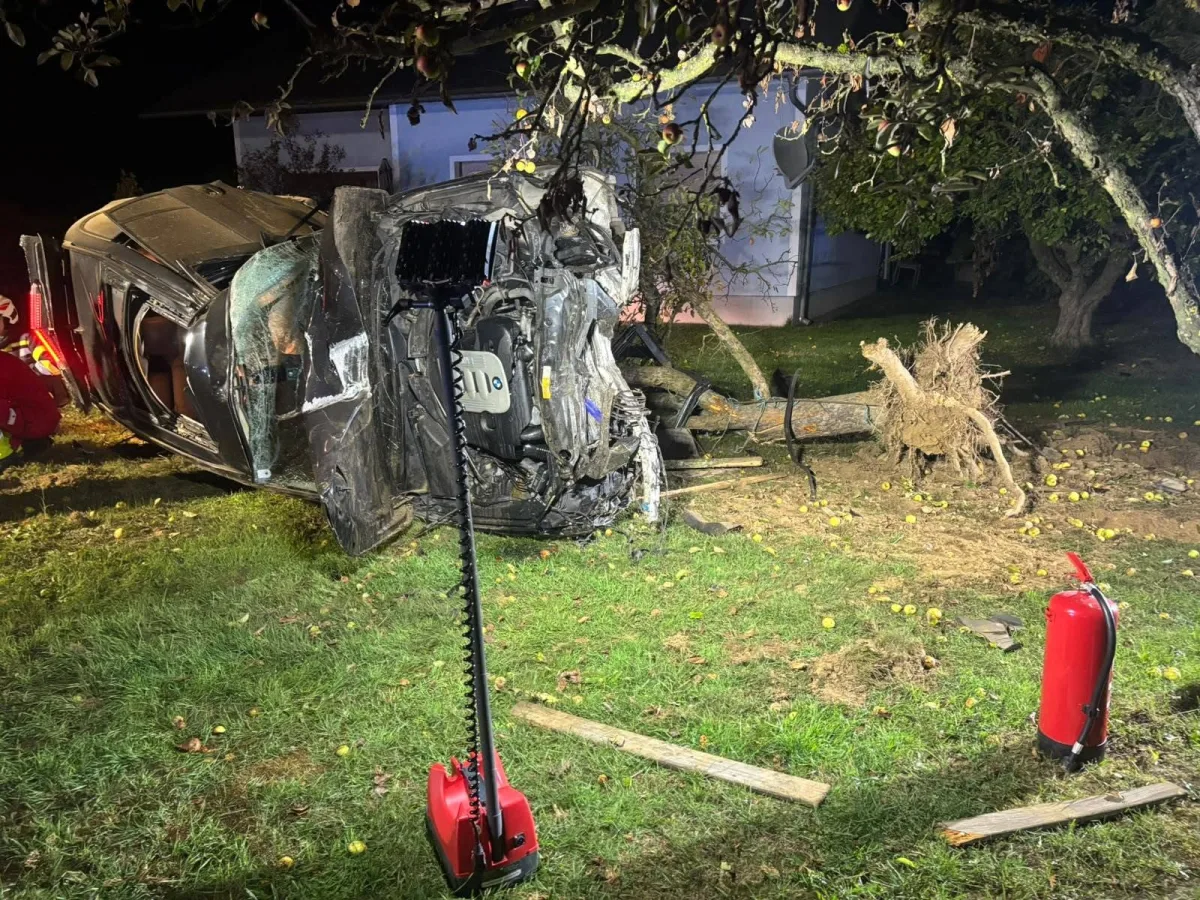 Ein Auto liegt bei Nacht seitlich auf dem Rasen, ein entwurzelter Baum ist daneben sichtbar. Im Vordergrund stehen zwei Feuerlöscher.