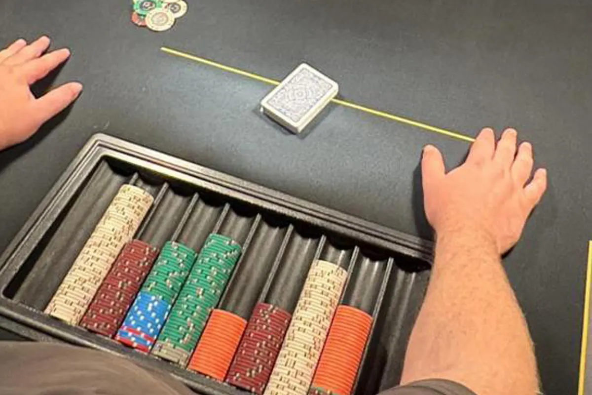 Auf einem Tisch befinden sich Pokerchips und ein Kartendeck. Zwei Hände liegen links und rechts vom Tisch.