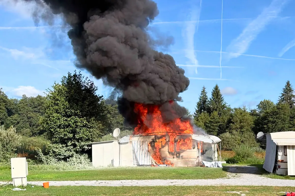 Ein Wohnmobil steht komplett in Flammen, dichter schwarzer Rauch steigt auf, umgeben von Bäumen und blauem Himmel.