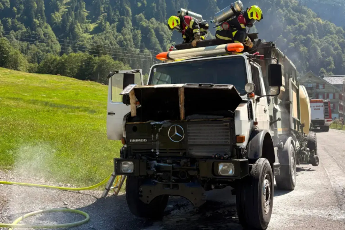 Zwei Feuerwehrleute auf einem Unimog-LKW, der auf einer Landstraße steht. Rauch steigt aus dem Motorraum auf.