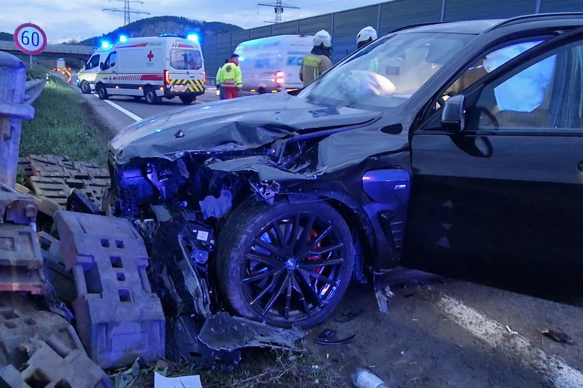 Ein stark beschädigtes schwarzes Auto steht neben der Autobahn. Im Hintergrund sind Notfallfahrzeuge mit Blaulicht und Personen in Warnkleidung zu sehen.