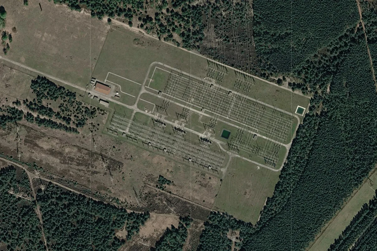 Die Aufnahme zeigt eine weitläufige Industrieanlage mit mehreren Gebäuden, umgeben von Wald. Das Gelände ist symmetrisch strukturiert mit Zufahrtswegen und Grünflächen.