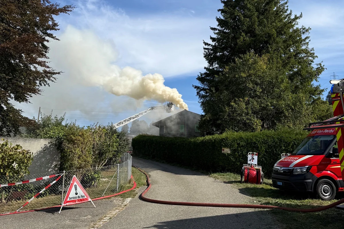 Rauch steigt aus dem Dach eines Hauses auf, während eine Feuerwehrleiter zur Brandbekämpfung eingesetzt wird. Ein Feuerwehrfahrzeug steht bereit, um die Löscharbeiten zu unterstützen.