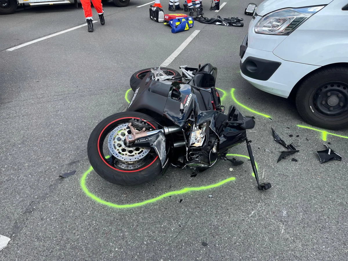 Ein Motorrad liegt beschädigt auf der Straße neben einem weißen Van, umgeben von Unfallmarkierungen und Trümmern.