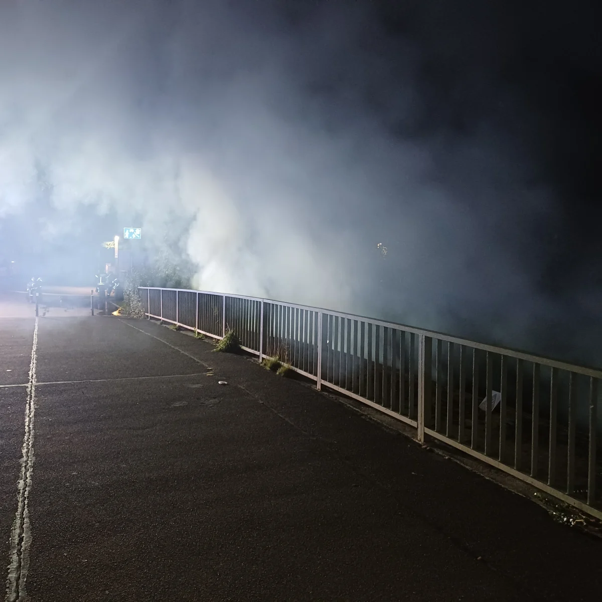 Eine Straße ist von dichtem Rauch eingehüllt, während Straßenlaternen die Szene bei Nacht erhellen. Ein Geländer verläuft entlang der Straße.