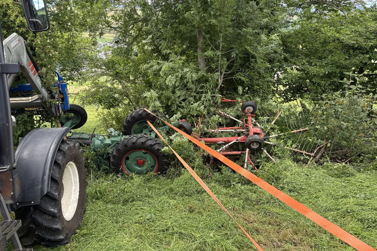 Ein Traktor ist mit einem Anhänger in einen Graben gerutscht und steckt zwischen Bäumen fest. Mehrere Spanngurte sind angebracht.