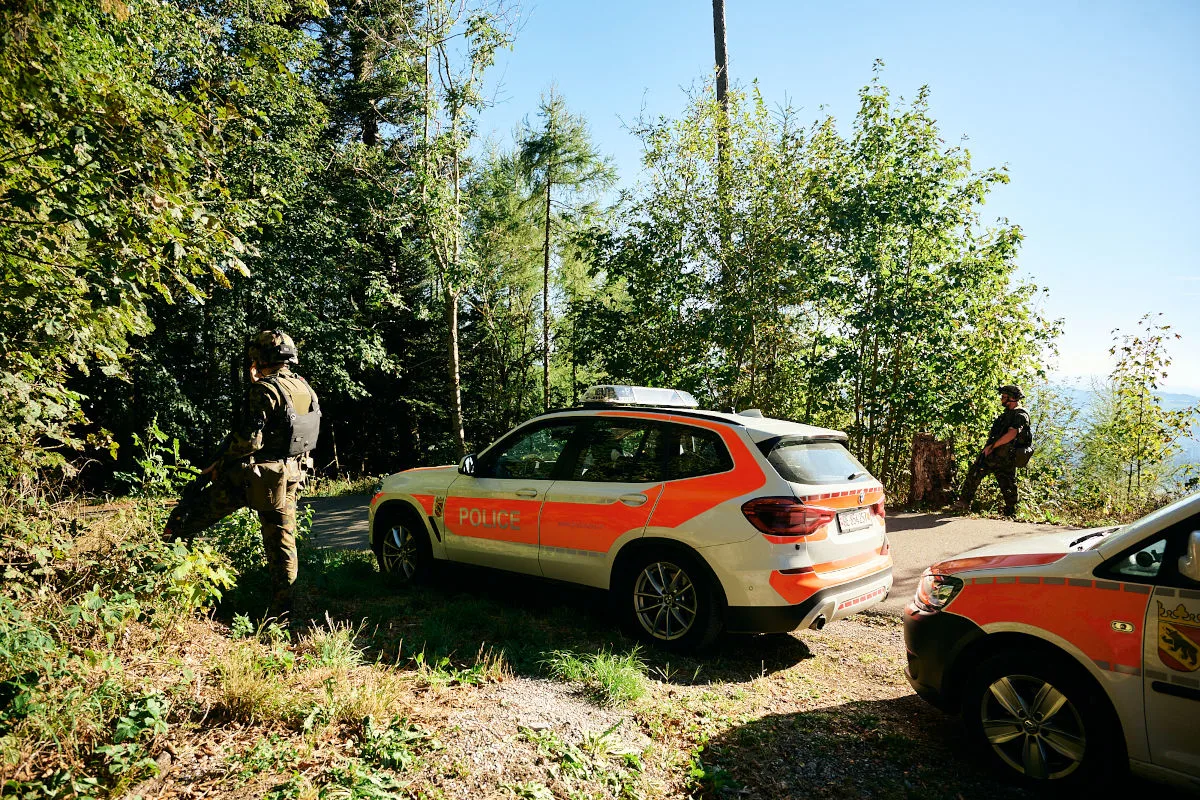Ein Schweizer Polizeiauto steht auf einer Waldstraße. Zwei Personen in Tarnkleidung sind links und rechts des Fahrzeugs sichtbar.