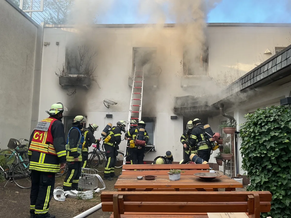 Mehrere Feuerwehrleute stehen vor einem Gebäude, aus dem dichter Rauch austritt. Eine Leiter führt zu einem oberen Fenster.