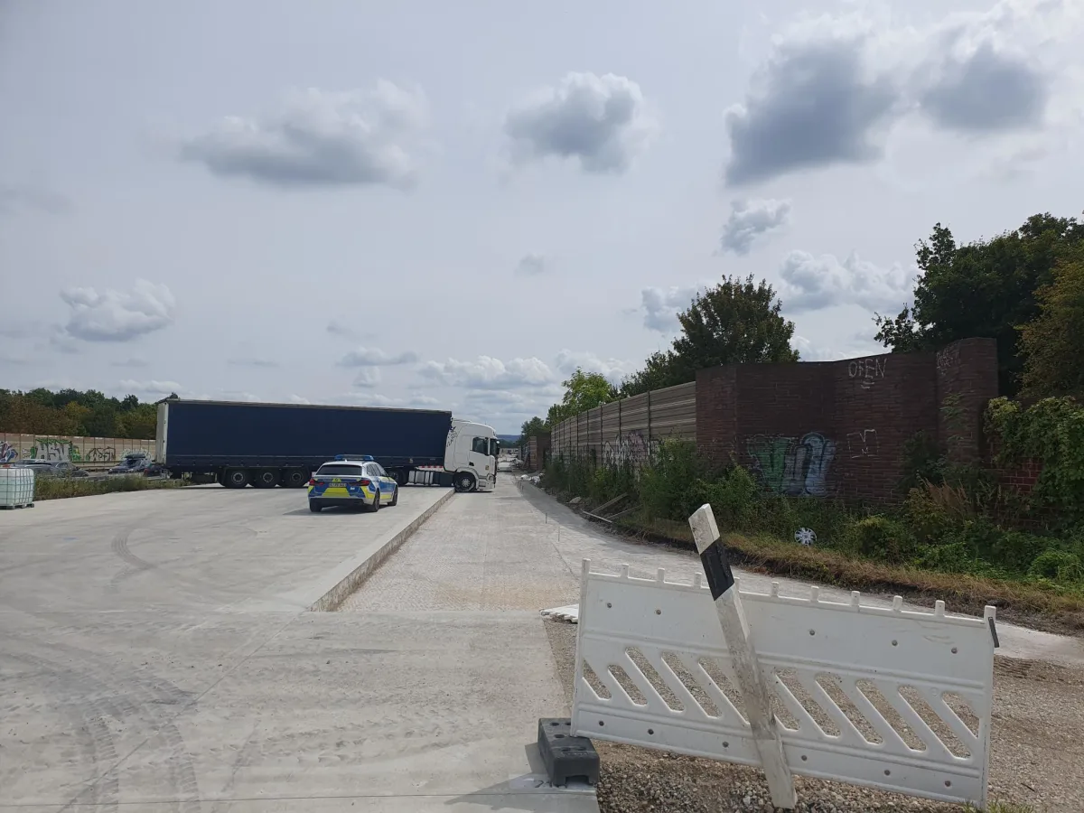 Ein Polizeiwagen parkt hinter einem großen Lastwagen auf einer asphaltierten Baustelle; im Hintergrund sind Bäume und Graffiti zu sehen.