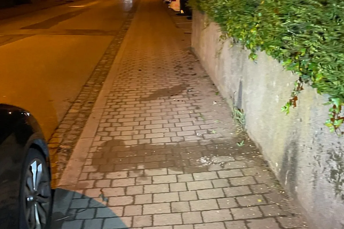 Ein beleuchteter Gehweg aus Pflastersteinen verläuft an einer Straße entlang. Links ist der Teil eines schwarzen Autos erkennbar.