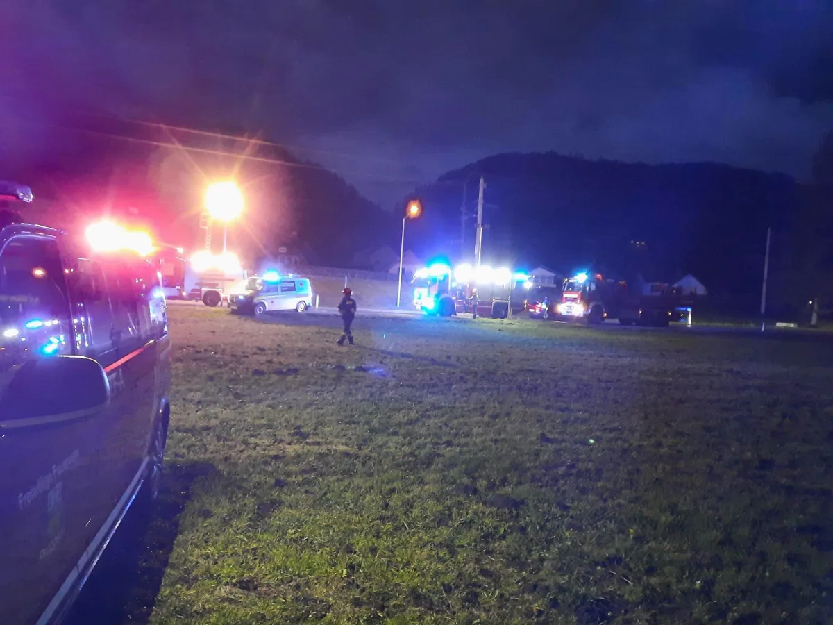 Bei Nacht sind mehrere Einsatzfahrzeuge mit Blaulicht auf einer Wiese zu sehen. Im Hintergrund erkennt man dunkle Hügel und vereinzelte Lichter.