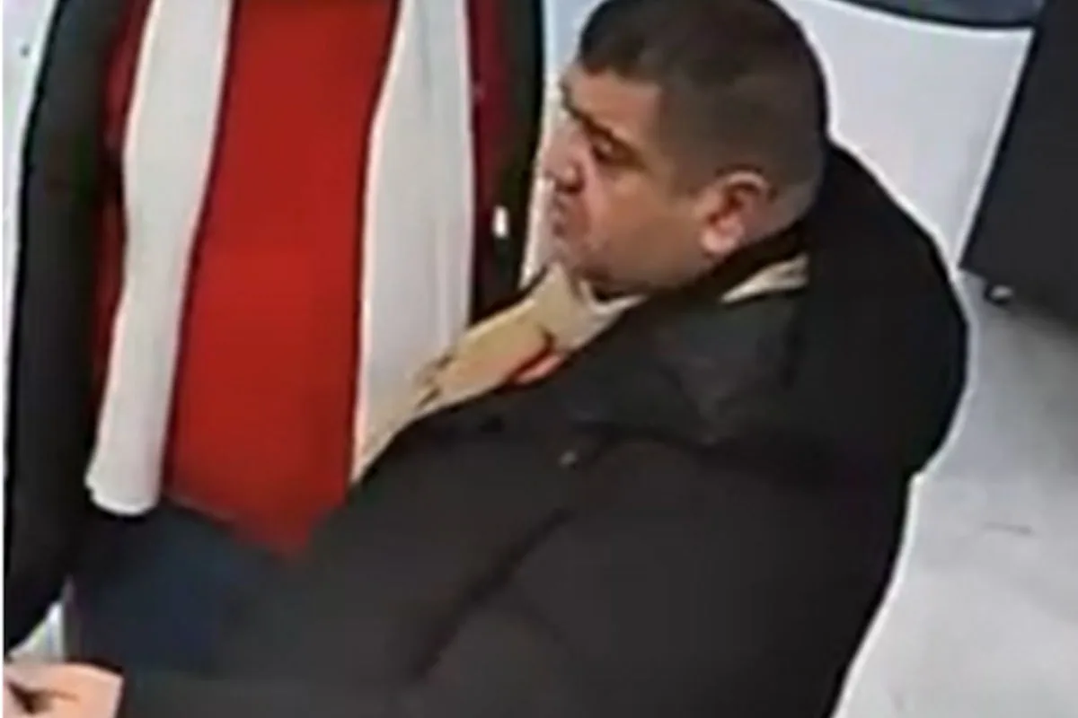 Ein Mann trägt eine dunkle Jacke und einen Schal und steht in einem geschlossenen Raum. Er scheint in ein Gespräch vertieft zu sein.