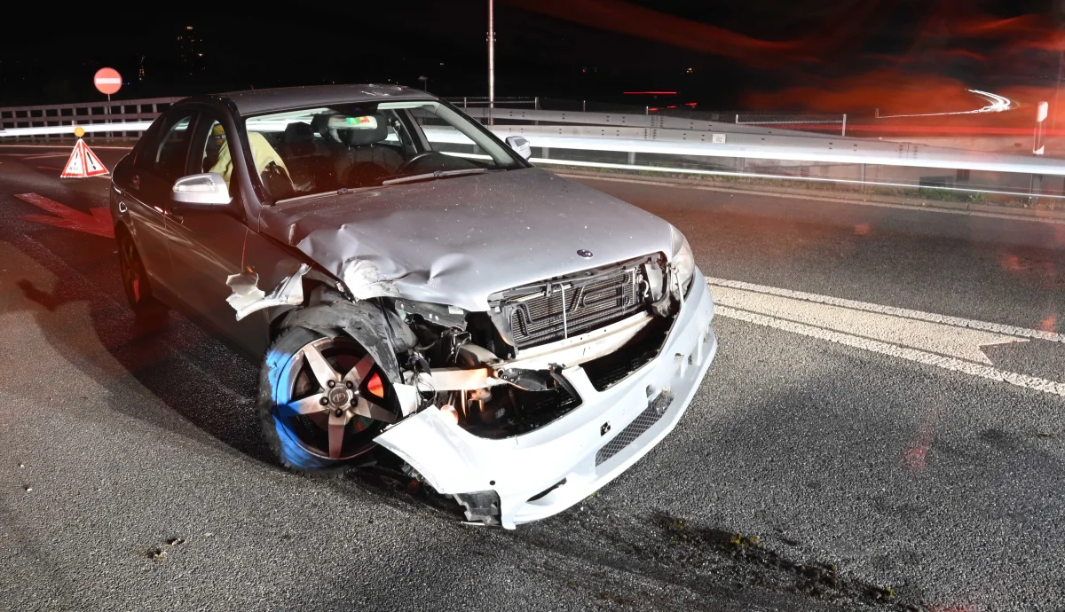 Ein silbernes Auto mit erheblichen Frontschäden steht auf einer nachtsbeleuchteten Straße. Im Hintergrund sind Leitplanken und Verkehrszeichen zu sehen.