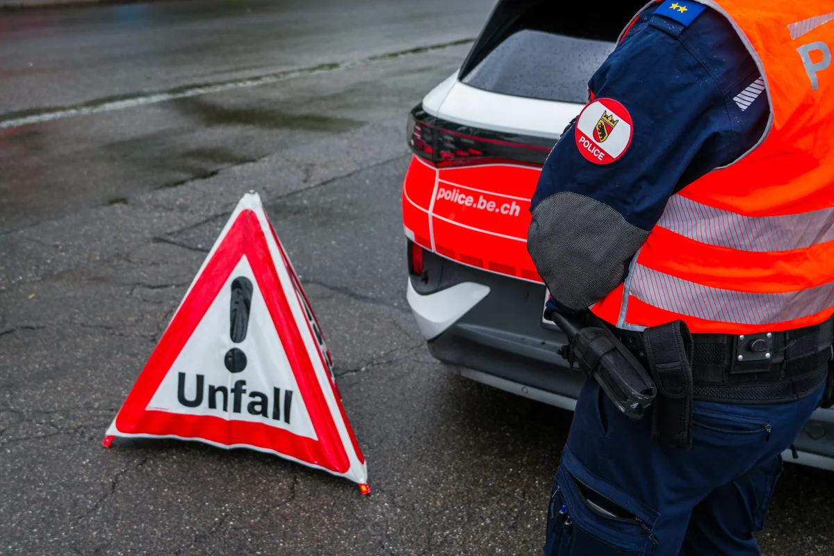Ein Polizist steht neben einem Unfallwarnschild auf einer feuchten Straße. Im Hintergrund ist ein Polizeifahrzeug sichtbar.