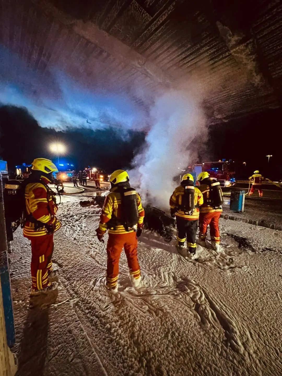 Mehrere Feuerwehrleute in voller Einsatzkleidung bekämpfen bei Nacht einen Brand unter einer Brücke mit starkem Rauch.