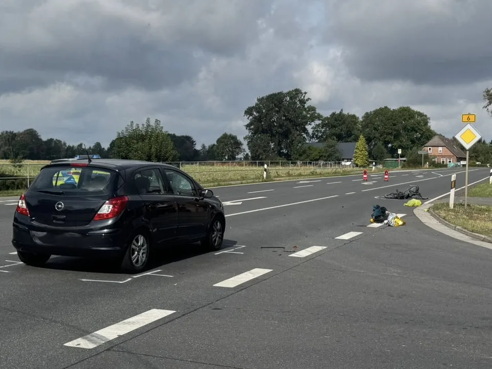Ein schwarzer PKW steht auf einer Landstraße, in der Nähe liegt ein umgestürztes Fahrrad und verstreute Gegenstände. Der Himmel zeigt eine Bewölkung.