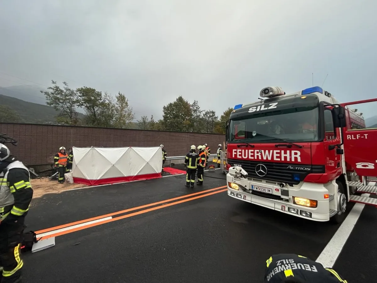 Ein rotes Feuerwehrauto der Marke Mercedes ist auf einer Straße zu sehen. Daneben stehen Einsatzkräfte neben einer weißen, mobilen Abschirmung.