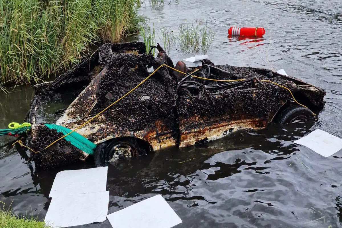 Ein stark verrostetes Auto liegt teilweise aus dem Wasser ragend in einem See. Es ist mit Seilen gesichert.
