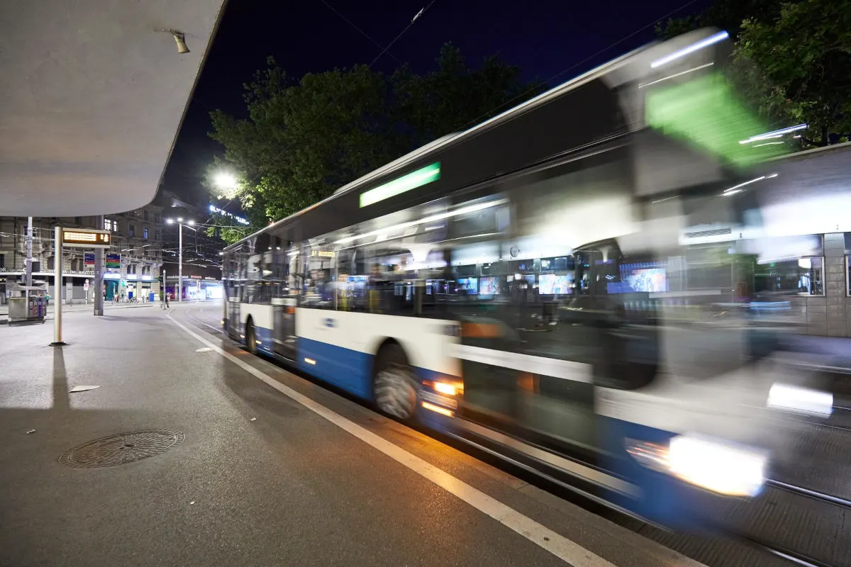Ein schnell fahrender Bus bewegt sich nachts entlang einer Bushaltestelle in einer beleuchteten Stadtumgebung.