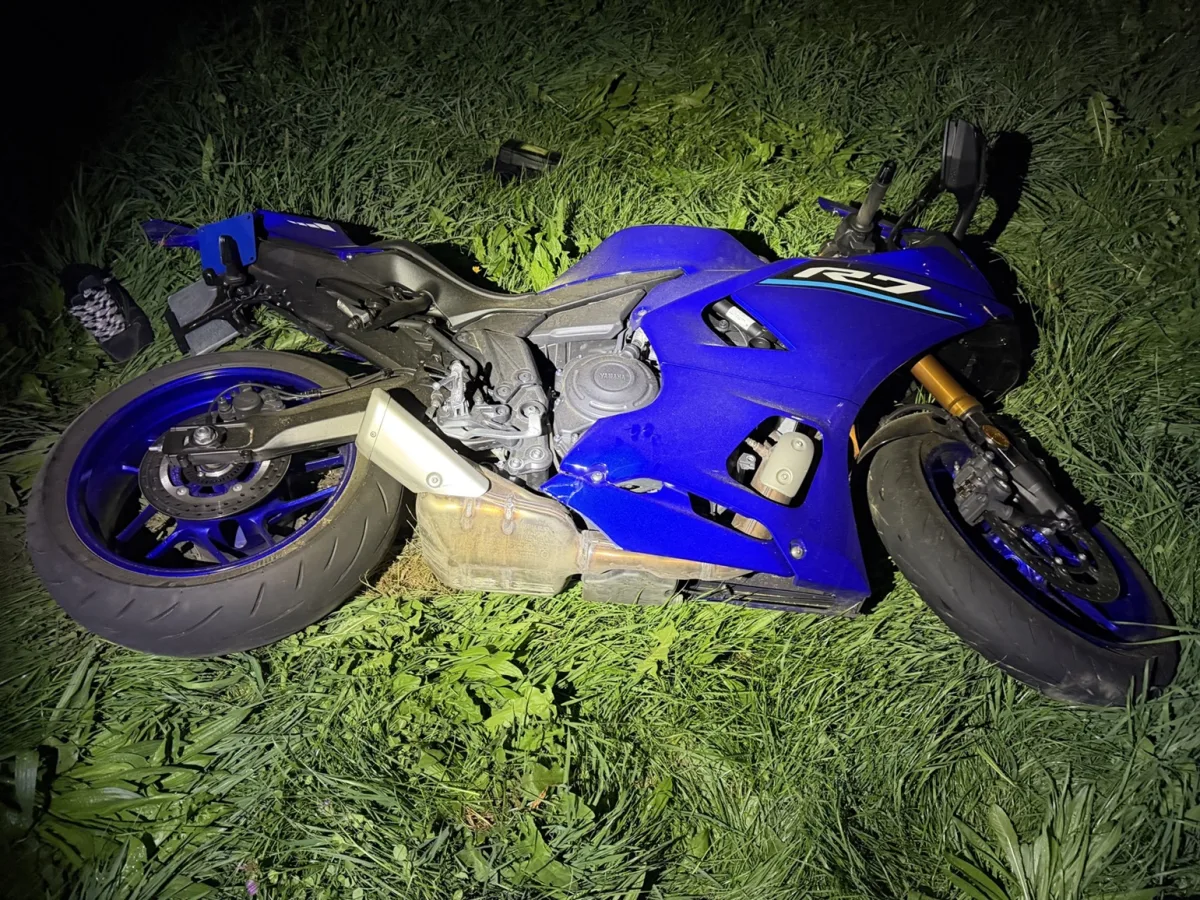 Ein blaues Motorrad liegt umgestürzt auf einer grasbewachsenen Fläche. Es ist Nacht, und das Bild wird durch künstliches Licht erhellt.