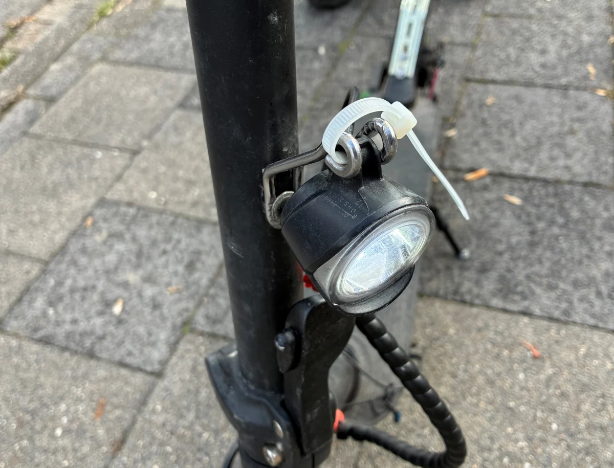 Nahaufnahme eines Elektroscooters auf einem Gehweg, an dem das Frontlicht mit einer Kabelbinder befestigt ist.