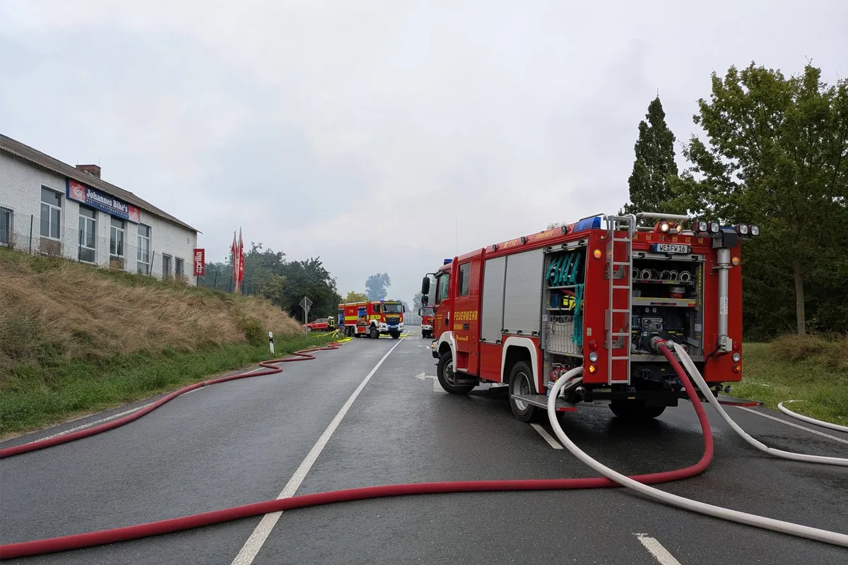 Mehrere Feuerwehrwagen stehen auf einer Landstraße, Schläuche sind ausgerollt und führen zu einem Gebäude neben der Straße.