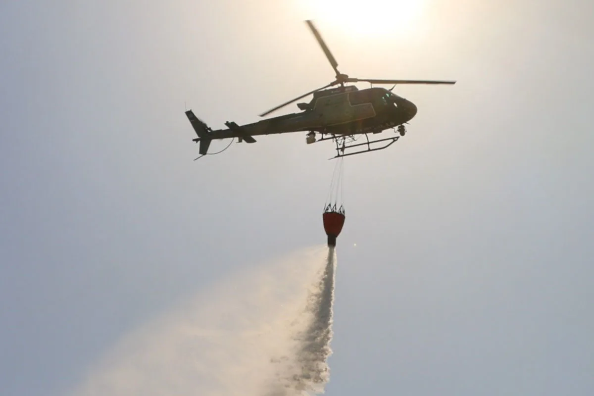 Ein Helikopter fliegt am Himmel und entleert einen großen, an einem Seil hängenden Wassertank, um einen Brand zu löschen.