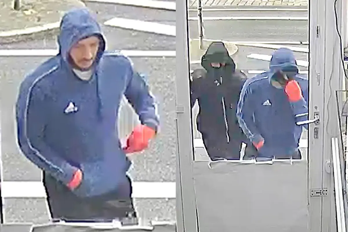 Zwei maskierte Personen mit Kapuzen, eine in blauem und eine in schwarzem Oberteil, betreten ein Gebäude durch eine Tür. Beide tragen Handschuhe.