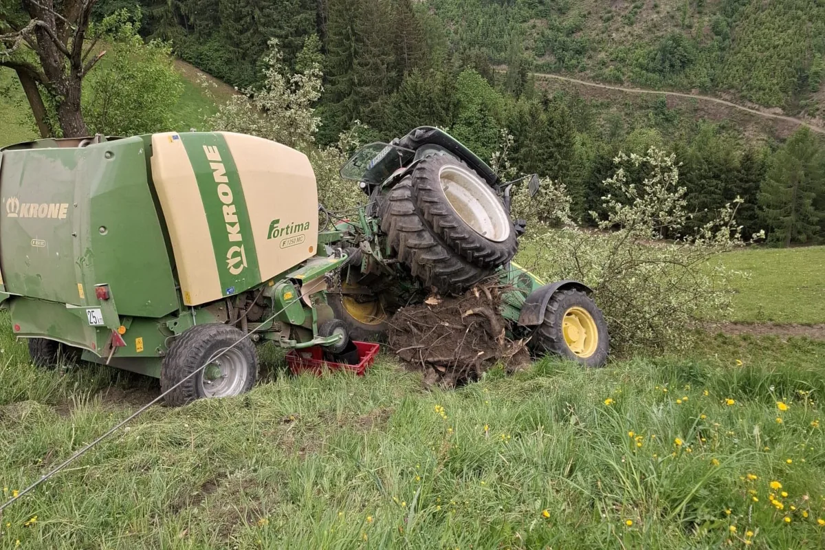 Ein grüner Traktor liegt umgestürzt auf einem Feld, umgeben von Bäumen und Gras. Im Hintergrund sind bewaldete Hügel zu sehen.