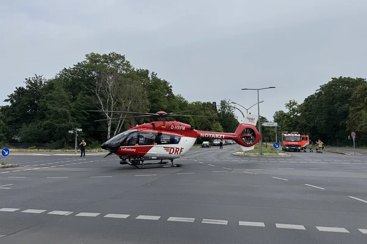 Ein DRF-Rettungshubschrauber steht auf einer breiten Straße, während im Hintergrund ein Feuerwehrfahrzeug und Einsatzkräfte sichtbar sind.