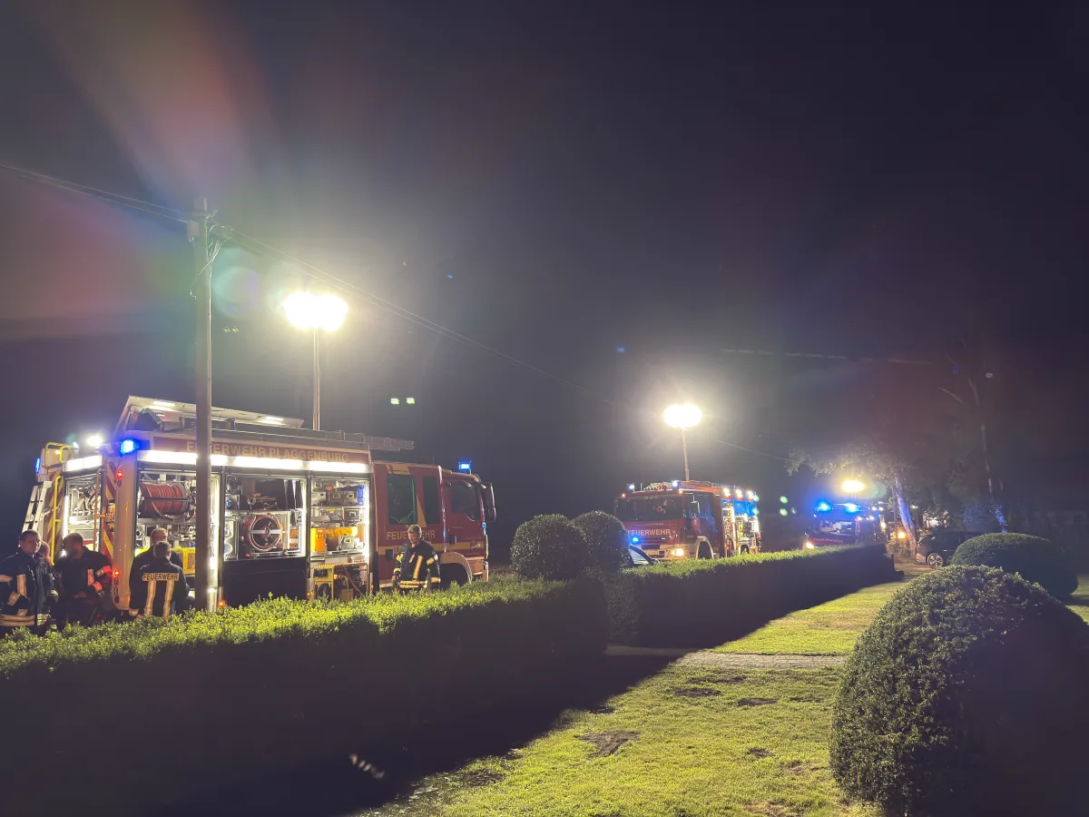 Zwei große Feuerwehrfahrzeuge stehen nebeneinander unter intensiver Beleuchtung in einer gepflegten, grünen Umgebung. Feuerwehrleute sind ebenfalls zu sehen.