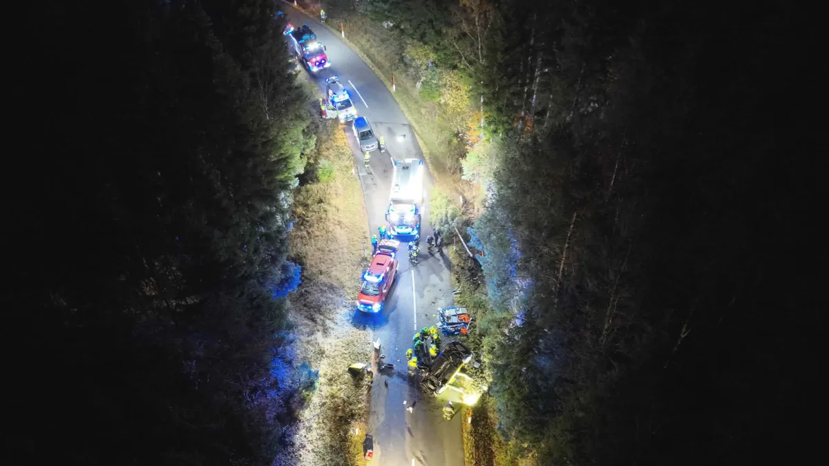 Mehrere Einsatzfahrzeuge und Feuerwehrleute sind auf einer engen, kurvigen Waldstraße zu sehen. Ein verunfalltes Auto liegt im Graben.