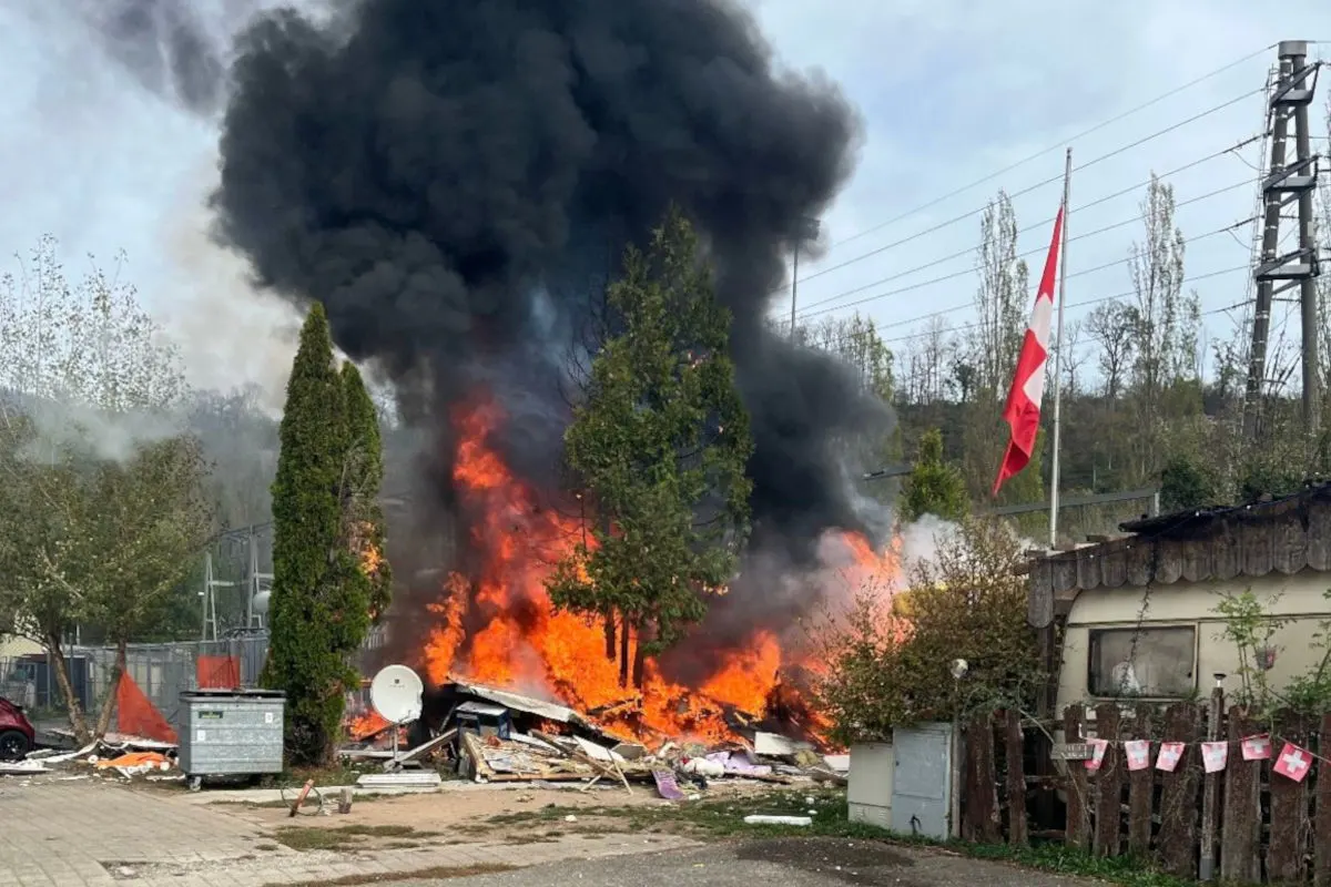 Ein intensives Feuer mit dicken Rauchwolken lodert in einer Wohnwagensiedlung. Eine Schweizer Flagge weht in der Nähe.